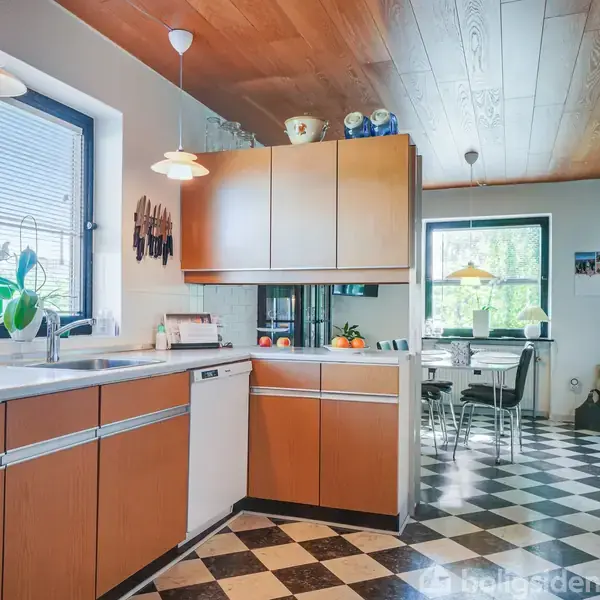 Et køkken med brune skabslåger og sort-hvidt ternet gulv. Bordet er fyldt med frugter og planter. I baggrunden er et spisebord ved et stort vindue.