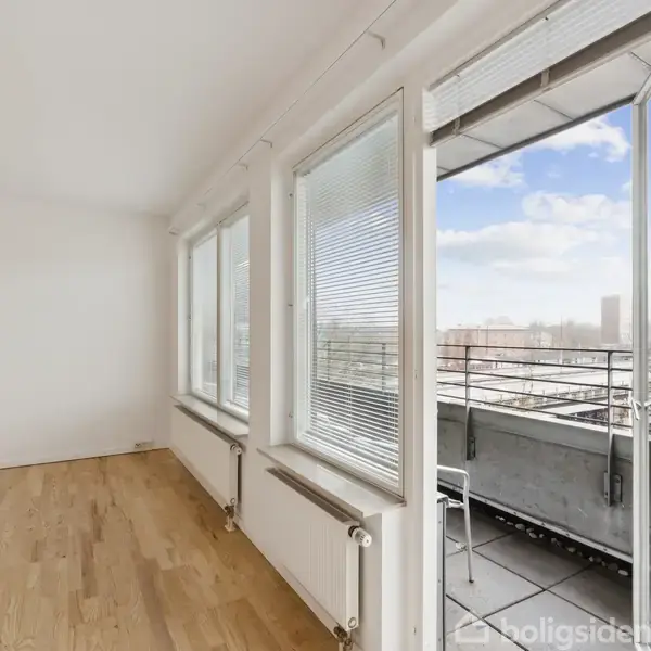 Et lyst, tomt rum med trægulve og hvide vægge. Et åbent køkken med en bardisk er placeret mod venstre baggrund. Til højre ses døre til andre rum.