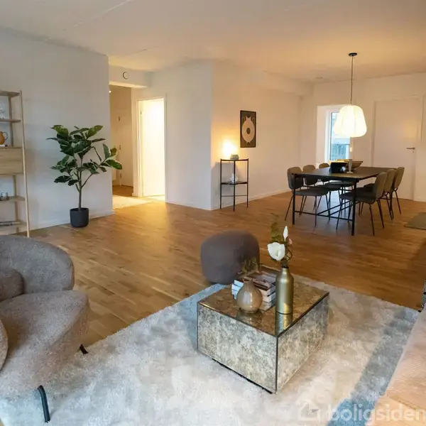 Stue med bløde lænestole og sofa omkring et sofabord med pynteting. Bagved står et spisebord med stole under en hængelampe. Rummet har trægulve og minimalistisk indretning.
