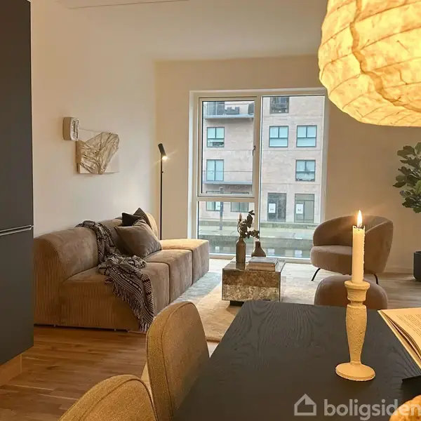 En stue med en beige sofa placeret mod en væg, en gulvlampe ved siden af, og et sofabord foran. Et dækket spisebord står i forgrunden, med tændte stearinlys. Store vinduer i baggrunden.