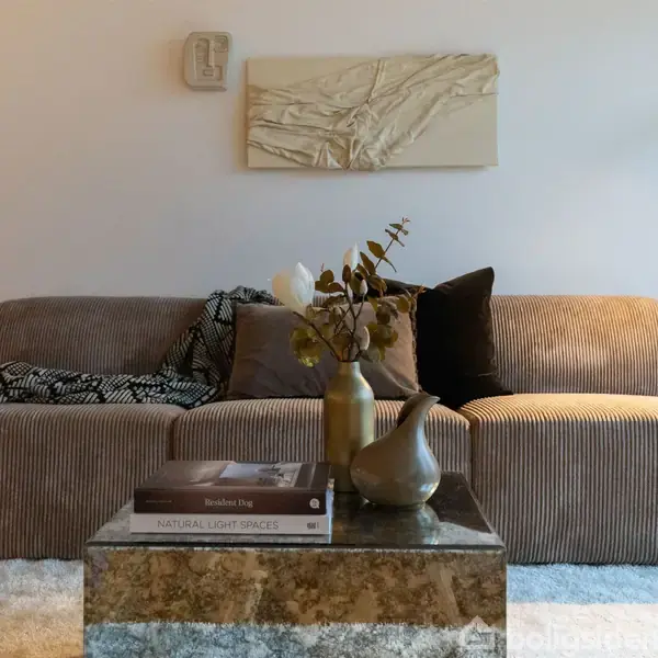 Brun sofa med stribemønster er pyntet med puder og tæppe. På sofabordet ligger bøger og en vase. En moderne gulvlampe oplyser rummet. Maleri og ur hænger på væggen.