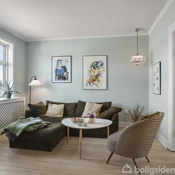 En stue med en brun sofa og et rundt sofabord ved siden af en mønstret stol. Væggene er lyseblå med to kunstbilleder, og store vinduer slipper lys ind.