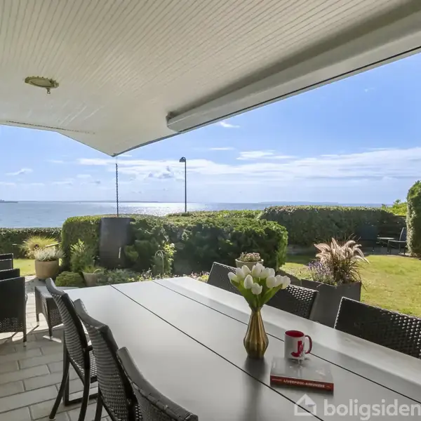 Terrassemøbler står omkring et langbord med en vase og kop ovenpå. Bagved ses en udsigt over en have og havet i baggrunden under en klar blå himmel.