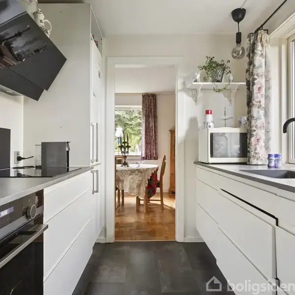 Køkken med hvide skabe og sorte bordplader; en ovn og kogeplade til venstre, en mikrobølgeovn og planter til højre. Udsigt mod en spisestue med spisebord og stole.