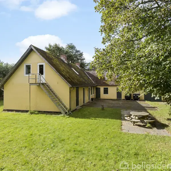 Et gult hus med en udendørs trappe placeret i græsplæne med picnicbord. Omgivet af træer under en klar blå himmel.