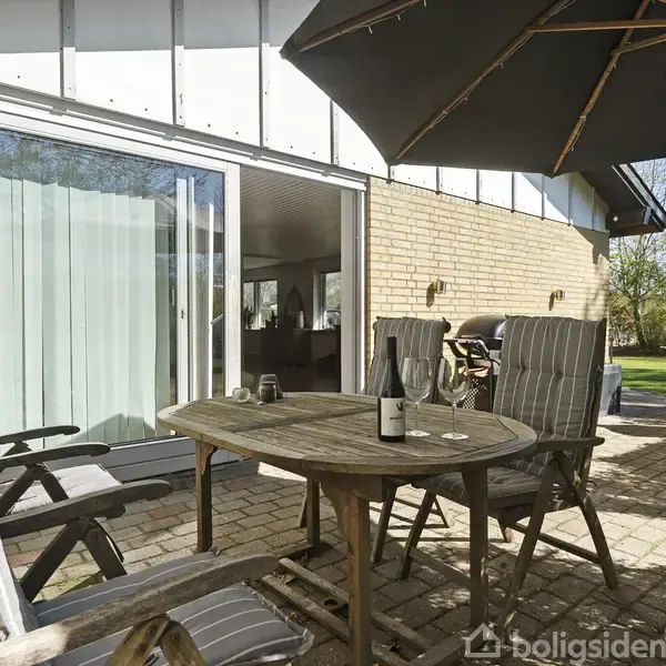 Et rundt havebord med stole står på en terrasse ved et hus med store glasdøre. En parasol og grill ses i baggrunden, mens en have strækker sig ud til højre.