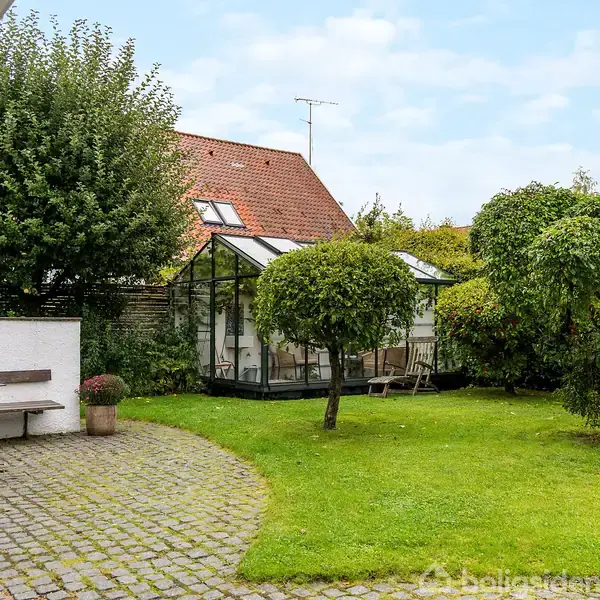 Drivhus står i have omgivet af græsplæne og træer. En brolagt terrasse med bænk og blomsterkrukke er til venstre. Hus i baggrunden med rødt tag.
