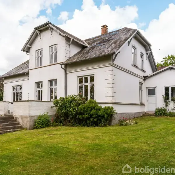 Hvidt hus står stille omgivet af træer og buske, med en grussti foran. Det ligger i landligt område med marker og skov i baggrunden.