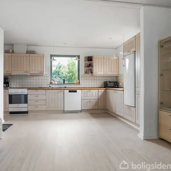 Et køkken med lyse træskabe er roligt indrettet, mens en opvaskemaskine står i midten. En hvid ovn og køleskab er placeret på hver sin side, med dagslys strømmer ind gennem et vindue.