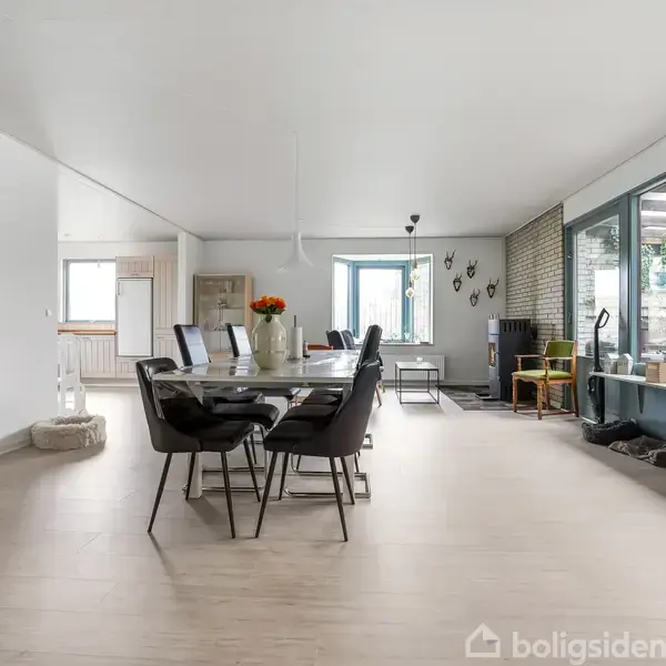 Et spisebord med sorte stole står i centrum. Bordet er pyntet med blomster. Rummet har lyse trægulve og store vinduer med udsigt til en terrasse.