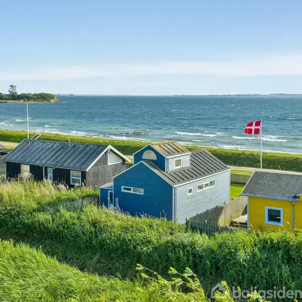 Farverige småhuse står på række ved havet. Det danske flag vajer i vinden, og grønt græs omkranser bygningerne. Den blå himmel og havet strækker sig i baggrunden.