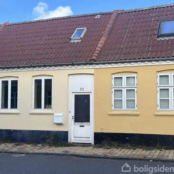 Et gult murstenshus med brune teglstenstage har en hvid dør med tallet "53". Vinduerne er forsynet med skodder. Huset ligger langs en asfalteret gade.