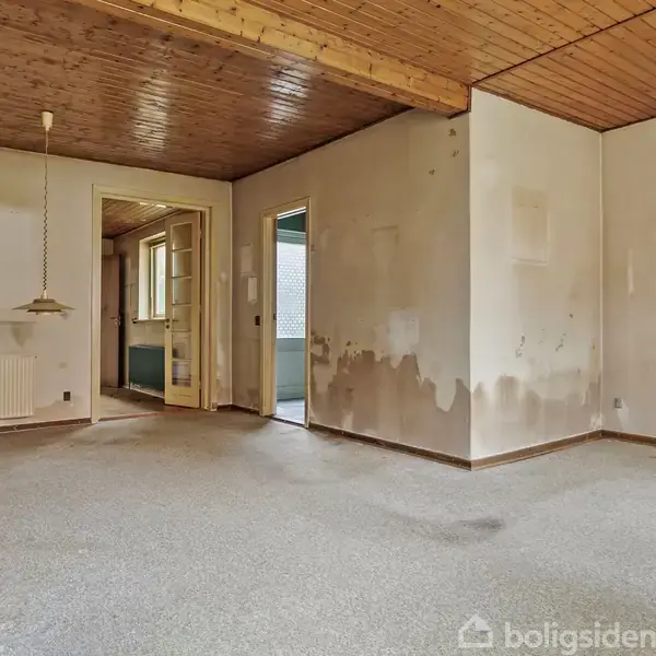 Tomt rum med pletvæggede vægge og træloft, indeholder en radiator og to hængelamper. Et åbent dørhul fører til en anden del af boligen.