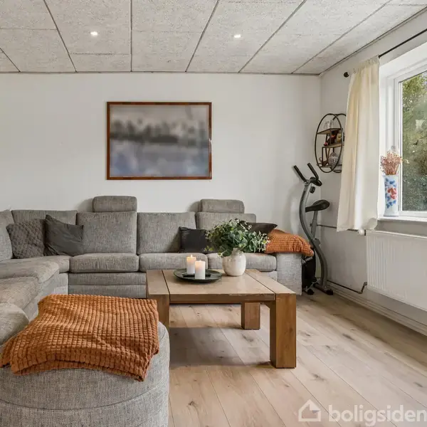 Grå hjørnesofa står mod en hvid væg med et maleri, omgivet af minimalistiske hylder. Dækket bord med planter og lys. Store vinduer giver udsigt til en grøn have.