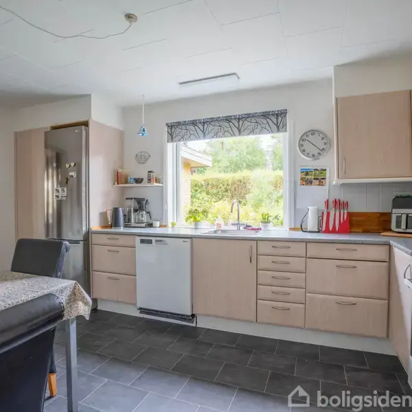 Køkken indeholder opvaskemaskine, komfur, køleskab og mikroovn, placeret i et lyst rum med vindue imod grønt udendørsområde. Bord med sorte stole står i forgrunden. En væg-ur viser tiden.