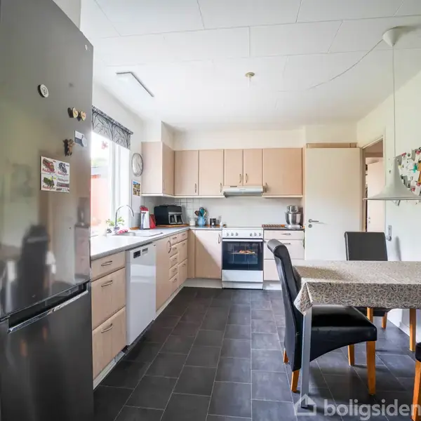 Køleskab står ved væggen, omgivet af køkkenskabe og en opvaskemaskine. Et bord med dækkeservietter og stole fylder rummet. Vindue lader lys ind i køkkenet.