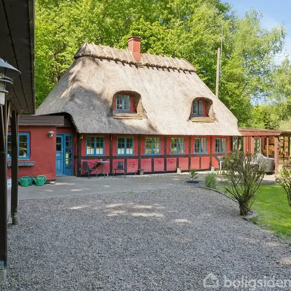 Et traditionelt bindingsværkshus med stråtag står stille omgivet af grussti og grøn vegetation. Husets facade er rød med hvidrammede vinduer, og en skov ses i baggrunden.