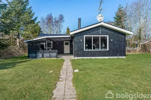 Et sortmalet hus står stille på en grøn græsplæne, omgivet af træer. En gangsti fører til hoveddøren, og satellitantenne ses på taget.