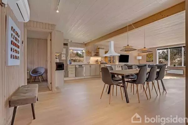 Et spisebord med otte stole står i et moderne åbent køkkenalrum. Baggrunden viser lyse trævægge, stort vindue med udsigt og hvid emhætte over køkkenområdet.