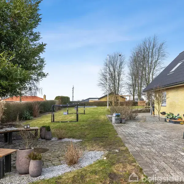 Gul hus med sort tag står ved siden af en stor have. Terrasse med fliser og havemøbler til højre, græsplæne og træer i baggrunden. Skiftende skyfri himmel.