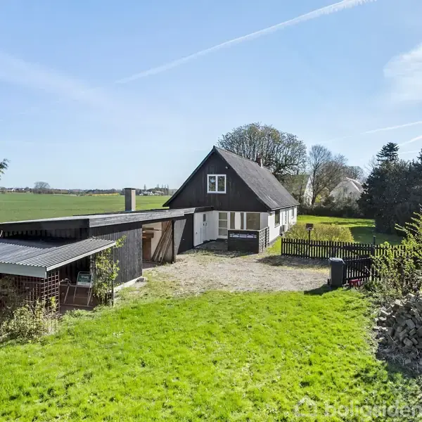 Et hvidt hus med sort tag ligger i en åben, grøn have. Der er en carport til venstre og marker i baggrunden under en blå himmel.