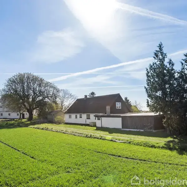 Hvidt, landligt hus med mørkt tag står stille i sollys, omgivet af grønne marker og store træer; skyer og flystriber på en klar blå himmel.