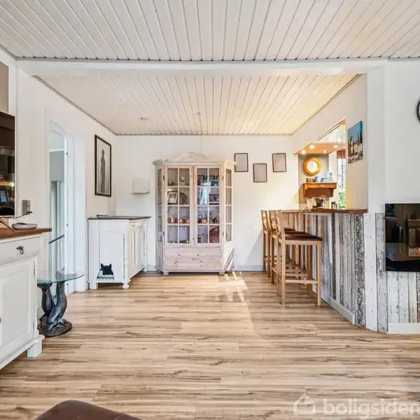 Et lyst stueområde med trægulv. En pejs er tændt ved væggen med en ur over. Et rundt bord med stole står nær en barområde.