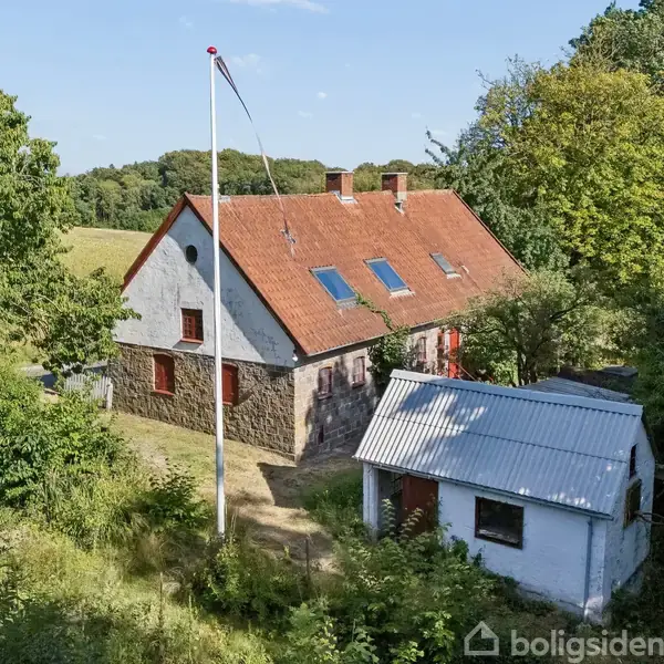 Stuehus med rødt tag står stille ved siden af en mindre, hvid bygning i en frodig, grøn landskab med træer og marker. En flagstang rejser sig foran huset.