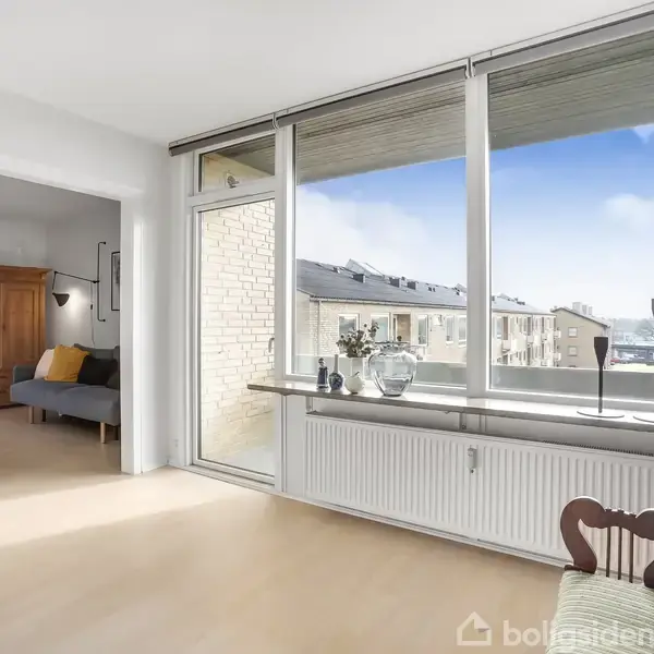 Et lyst rum med et klaver placeret ved et stort vindue. Vindueskarme har planter og lysestager. I baggrunden er en stue med sofa og skab synlige.