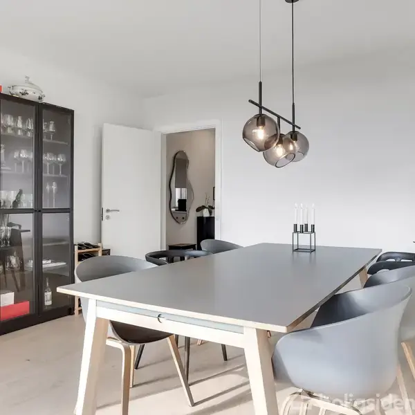 Et spisebord omgivet af grå stole står under tre hængende lamper. En glasvitrine fyldt med glasvarer står til venstre, og en grøn plante pryder højre side af rummet.