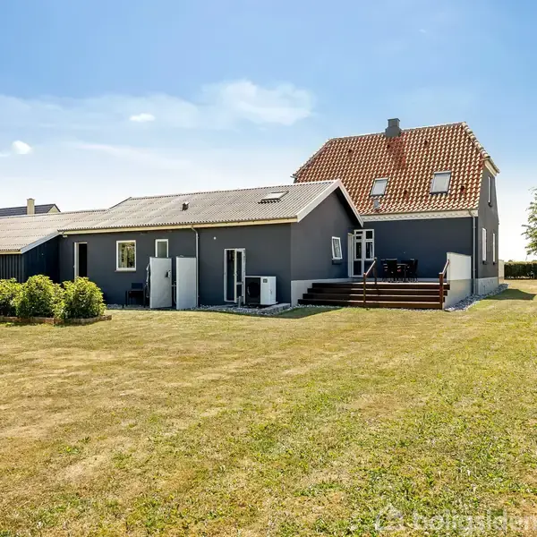 Huset står stille og er moderne med gråt eksteriør og rødt tegltag, omgivet af en stor, velplejet græsplæne under en klar blå himmel.