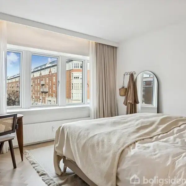 Soveværelse med dobbeltseng dækket af beige sengetøj. Et skrivebord og stol står ved stort vindue med udsigt til bygning. Gardiner omkranser vinduet. Bright og moderne indretning.