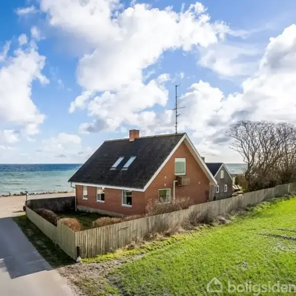 Et rødt murstenshus med sort tag står omgivet af hegn. Huset ligger tæt på havet, med græsmark til højre og en lavvandet sti foran, under en skyet blå himmel.