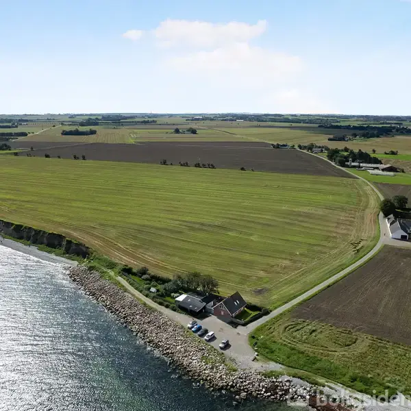 Landområde strækker sig langs kysten, med en vej, der snor sig gennem markerne. Flere huse ligger tæt på vejen. Vandet glitrer ved siden af kysten under en klar himmel.