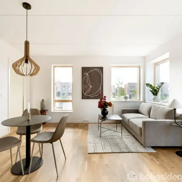 En stue med sofa står ved siden af et sofabord med en vase. Rundt spisebord med tre stole. Rummets lysindfald kommer fra tre vinduer, omgivet af hvide vægge.