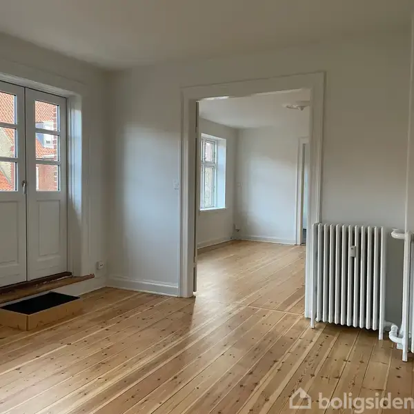 Dør stående åben, leder ind til rum med trægulv og hvide vægge; en radiator står ved siden af dørkarmen. Belysning fra vinduer i baggrunden skaber et lyst miljø.