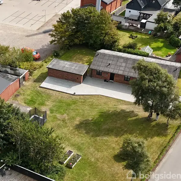 En rød murstensbygning ligger ved en vej, omringet af grønne træer og en velholdt græsplæne. Omkring huset er der yderligere bygninger og parkeringsområder.