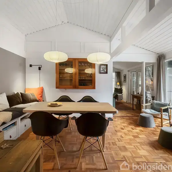 Et moderne spisebord med sorte stole står i en stue med hvid malet træloft. Rummet har store vinduer, lys indretning og to hængende lamper over bordet. To tændte lys kan ses til venstre.