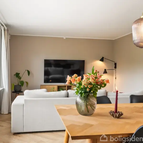 Sofa står foran vægmonteret TV i lys stue med træbord og vase med blomster, omgivet af store vinduer og hængende lampe. To stearinlys og en plante står på bordet.