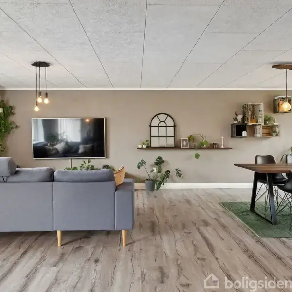 En stue med grå sofaer mod en beige væg, hvor et fladskærms-TV hænger. Til højre står et spisebord med stole på et grønt tæppe. Planter og lamper tilføjer dekoration.