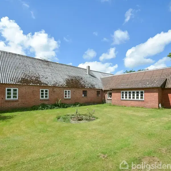 En gammeldags murstensgård med skråt tag står omgivet af en grøn græsplæne under en skyfri blå himmel. Der er træer på begge sider af bygningen.