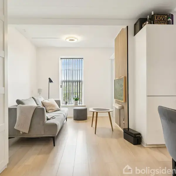 Sofa står ved siden af et gulv-til-loft vindue, omgivet af hvidmalede vægge. Til venstre ses en standerlampe; til højre et køkkenområde med skabe. Ingen tekst registreret.