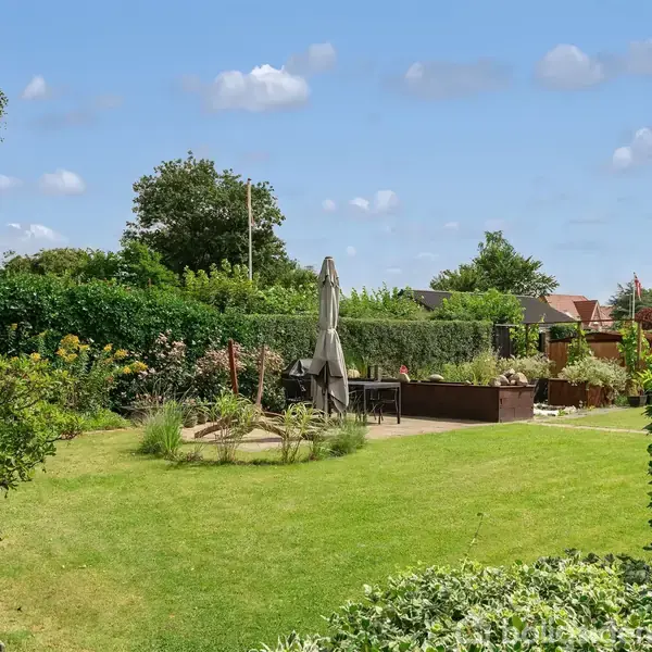 En velplejet have med en grøn plæne og tætte buske omkranser et haveområde med havemøbler og en stor parasol. Blå himmel med få skyer danner baggrund.