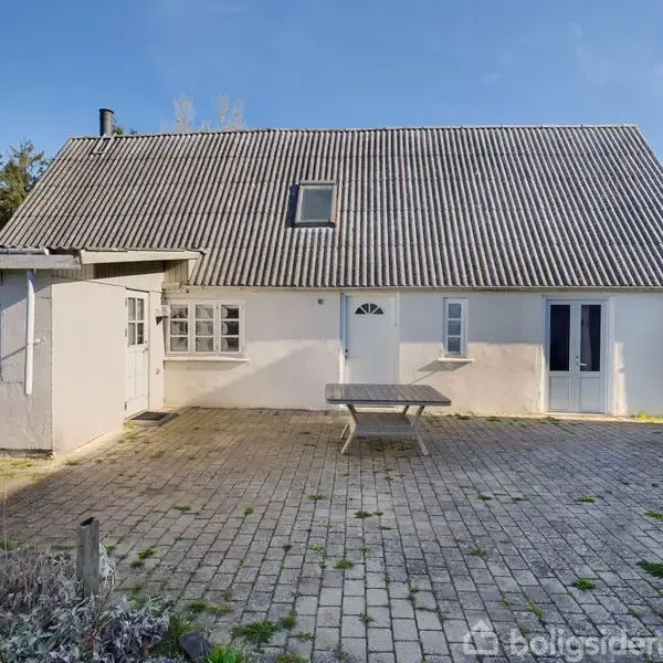 Hus med hvid facade og gråtag står på en gårdsplads af fliser. Et bord er placeret i midten. Træer og blå himmel ses i baggrunden.