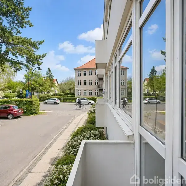 En cyklist kører på en vej foran en bygning med rødt tag omgivet af biler og træer. Forgrund viser en hvid bygning med store vinduer.