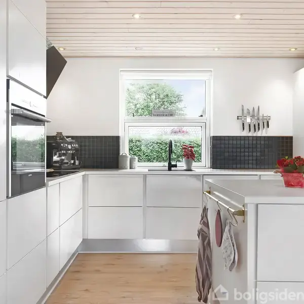 Et lyst, moderne køkken med hvide skabe og sort stænkpanel. En ovn indbygget i skabene. Et vindue med udsigt til træer og grønne planter. En kande og en plante står på bordet.