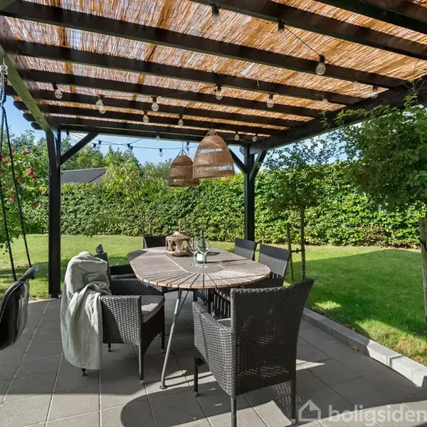 Pergola med flettag over ovalt træbord og seks kurvestole. Havegyngestol hænger i hjørnet. Grønt græs og tætte buske omkranser området. To lampekurve hænger over bordet.