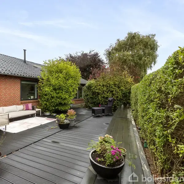 Terrassebord i træ med blomsterpotter på, omkranset af grøn hæk og murstenshus med vindue. Sofa og havestole placeret langs væggen i en afslappet, frodig haveindstilling.
