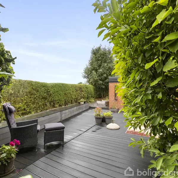 Terrasse med sorte trægulve og en grå lænestol. Planter og buske omkranser området. En lille murstensvæg er synlig i baggrunden, mens krukker med blomster står ved stolen.