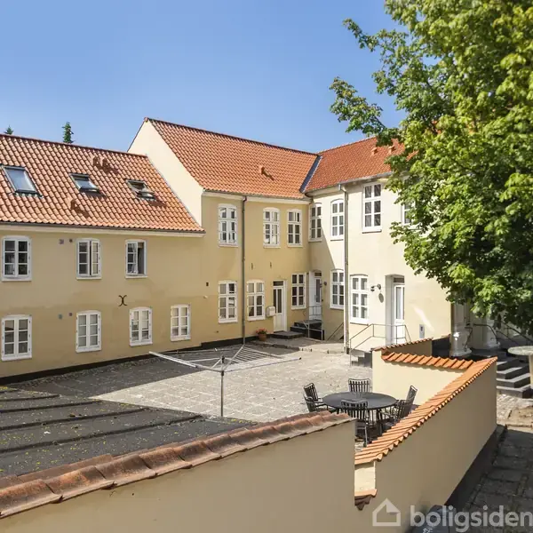 Gul bygning med røde tegltage omgiver en flisebelagt gård med siddepladser og parasoller. Omgivet af grønne træer under en klar blå himmel.